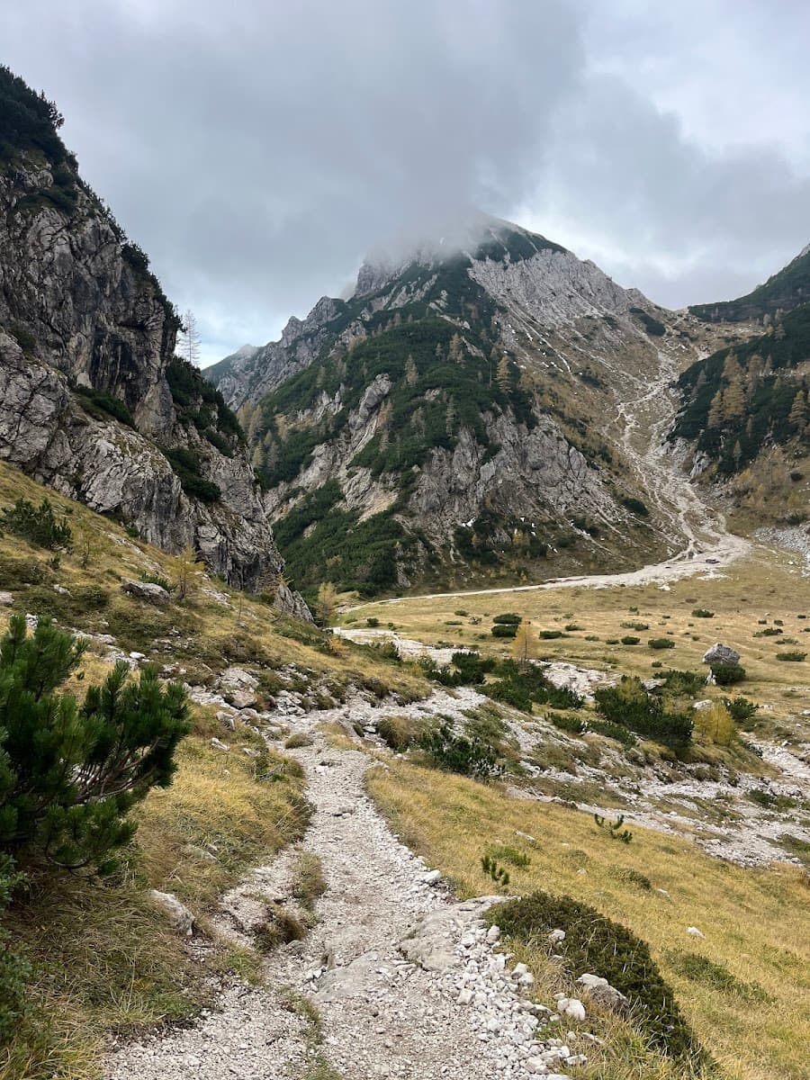 Rocky hiking trail leading into a valley between steep, forested mountains under a cloudy sky.
