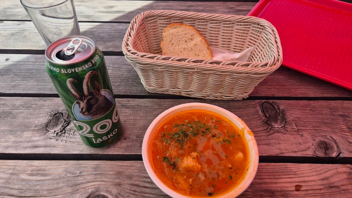 Bowl of orange soup, bread basket, and Laško beer can on wooden table
