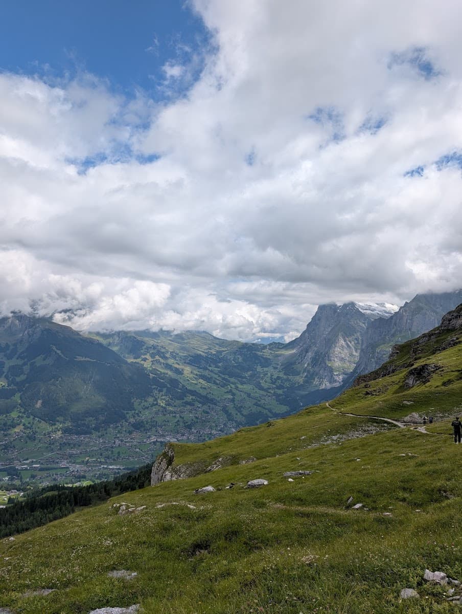 Hiking trail on grassy slope overlooking green valley and steep mountains under cloudy sky