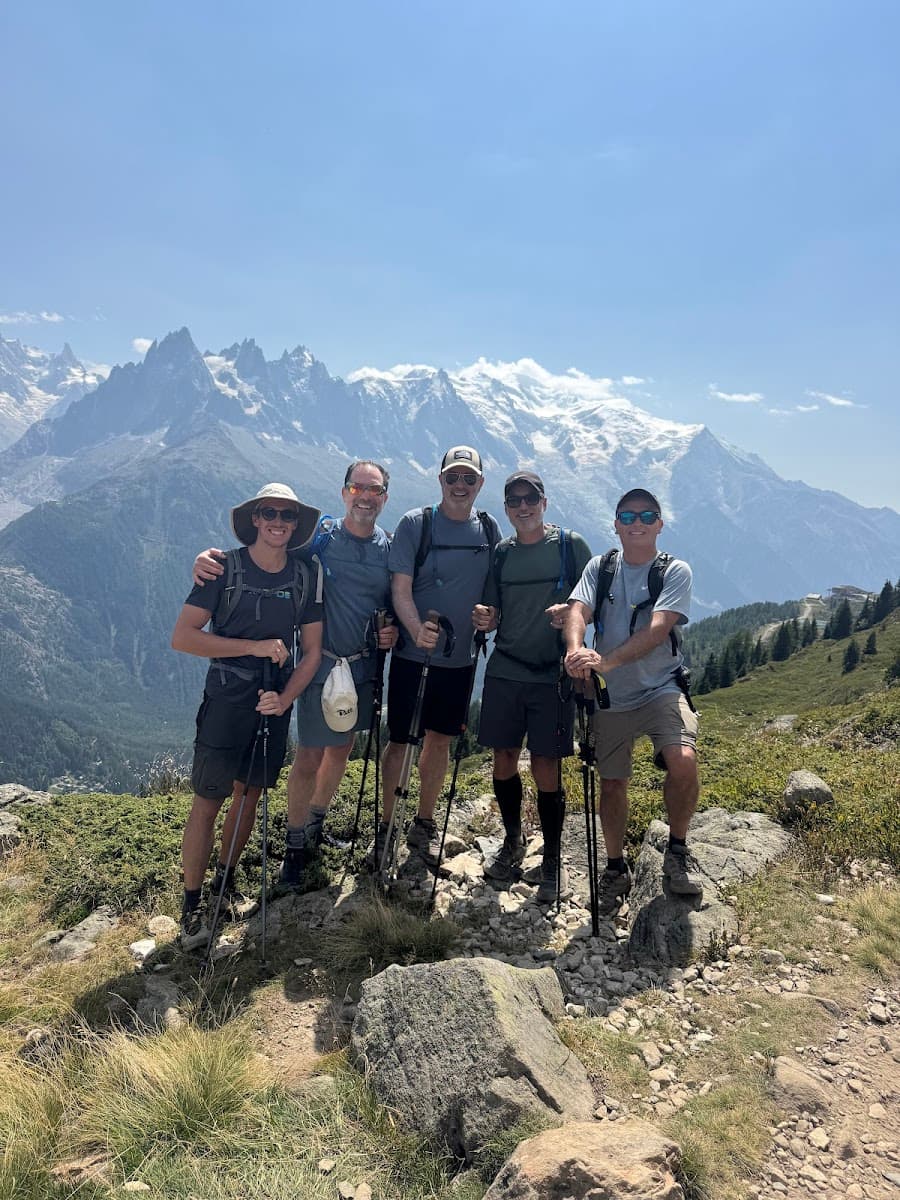 Hikers with trekking poles posing on rocky alpine trail with snow-capped mountains.