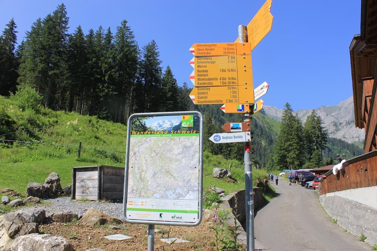 Hiking trail map and yellow signpost in a green alpine setting with mountains and forest