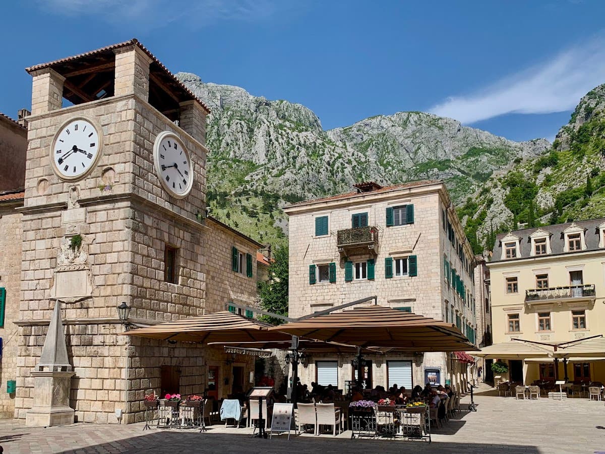 Stone clock tower and outdoor cafe dining in a square beneath steep, rocky mountains.