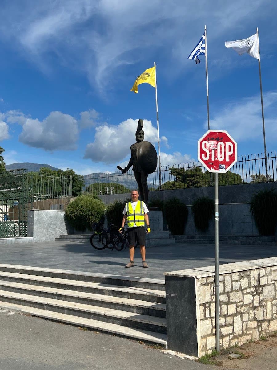 Cyclist standing by Spartan statue with Greek flag under blue sky