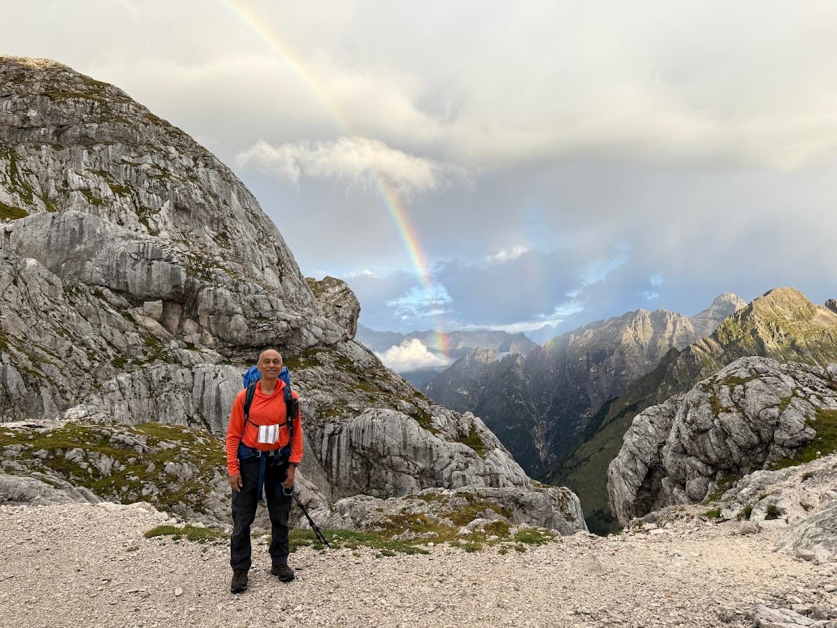 Hiker with backpack standing on rocky trail beneath rainbow over rugged mountains
